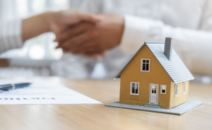 Miniature yellow house next to a contract with two people shaking hands in agreement